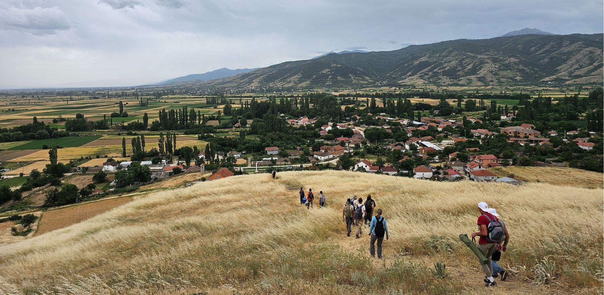 People walking across a grassy hillside, with a valley, houses, trees, and mountains in the background