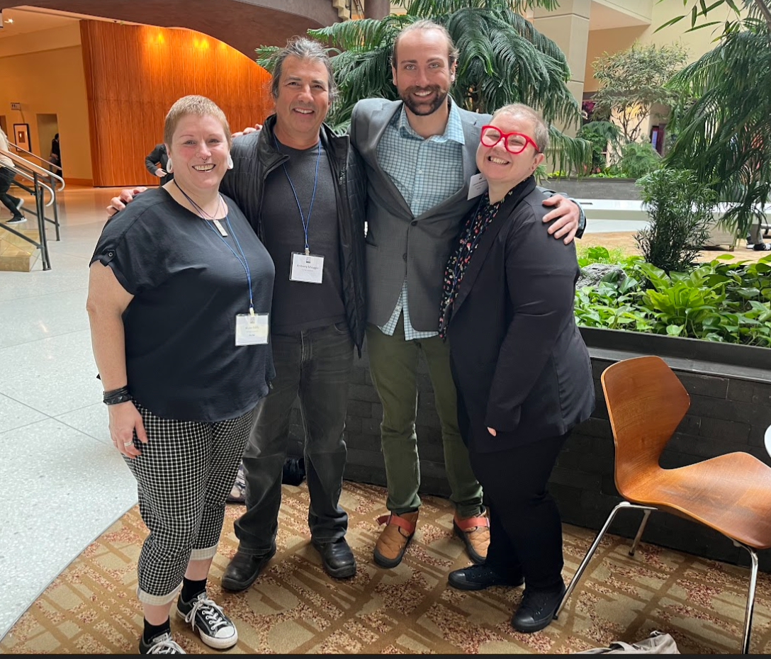 three alumni and one professor standing together smiling for the camer in a hotel lobby