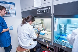 One student sitting in front of lab equipment wearing a white lab coat and a person with a blue lab jacket watching