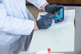 Hands wearing black latex gloves holding a device in front of a lab book.