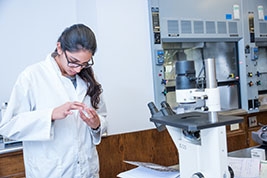 A student with a white lab coat identifying something and standing in front of a microscope