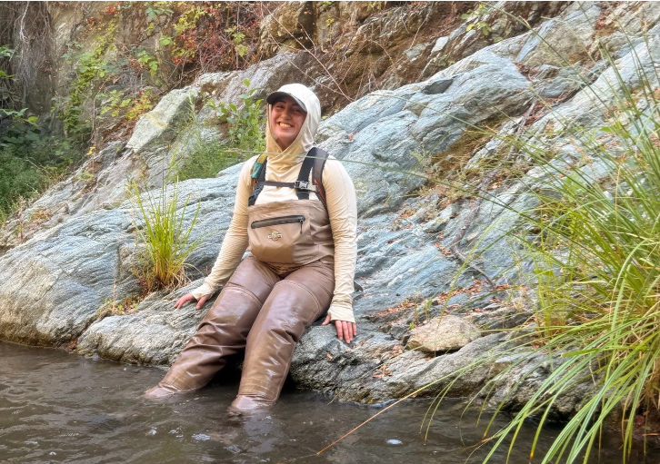 A student wearing waders, long sleeves, and a hat smiles at the camera while resting against a large rocky outcrop with their feet submerged in a flowing creek.