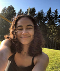 cami taking a selfie, wearing a black tank top, sitting on grass with trees in the background
