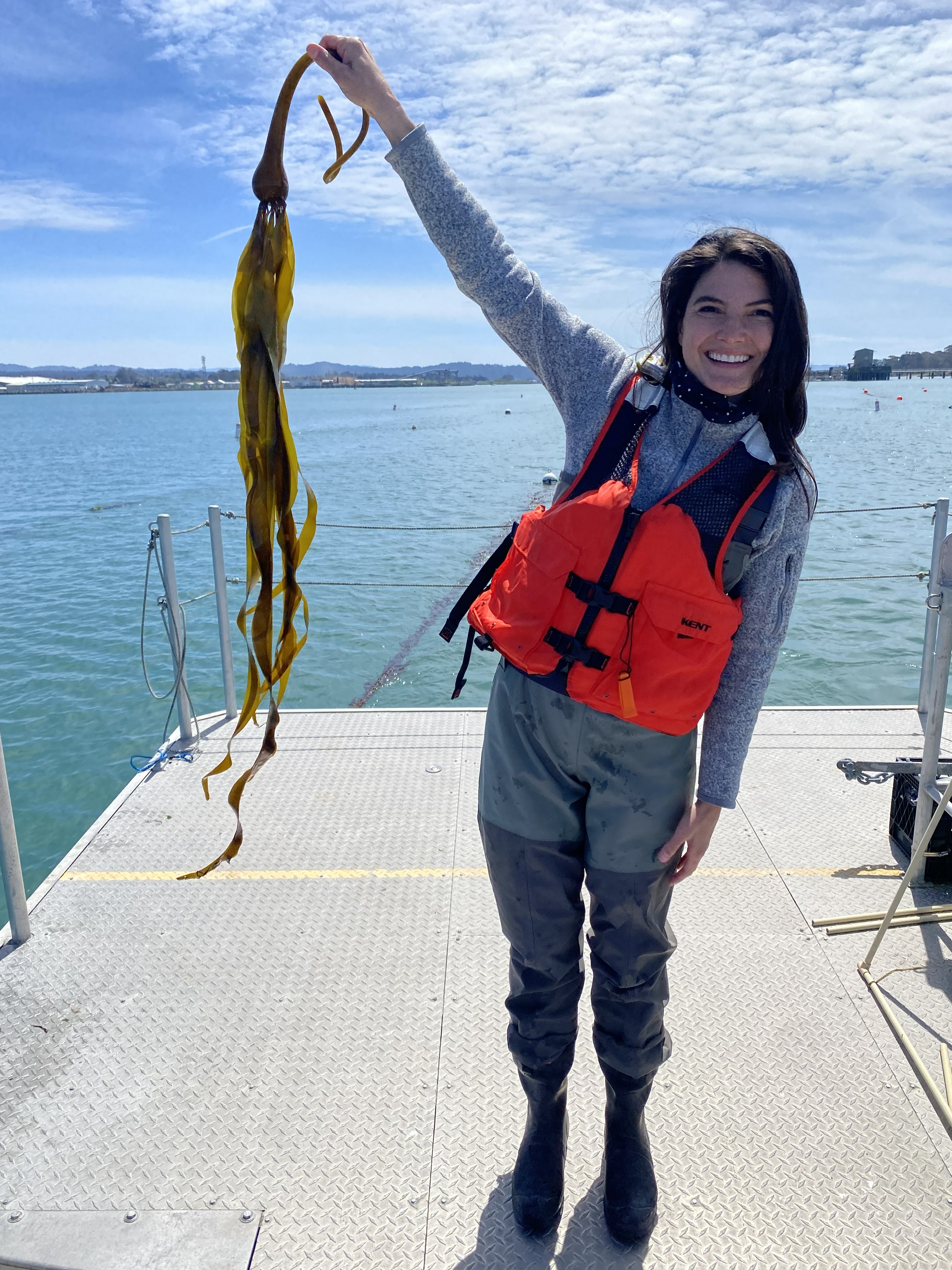 Jody Martinez standing on a peir over water. In her right hand she is holding up a strong of kelp, smiling with her life vest on.