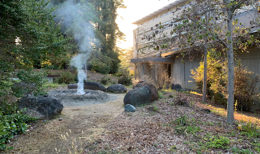Salmon Cooking Pit being used, smoke lifting above the put with the Native American Forum on the right side.