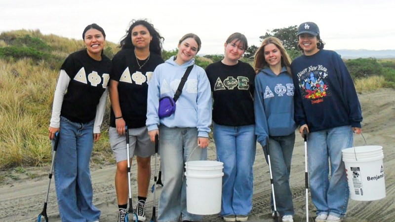 Six smiling Delta Phi Epsilon club members with buckets and trash pickers at the beach.