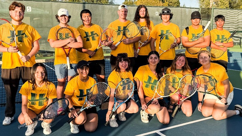 A team photo of the Humboldt tennis club in yellow shirts holding rackets on the tennis court.