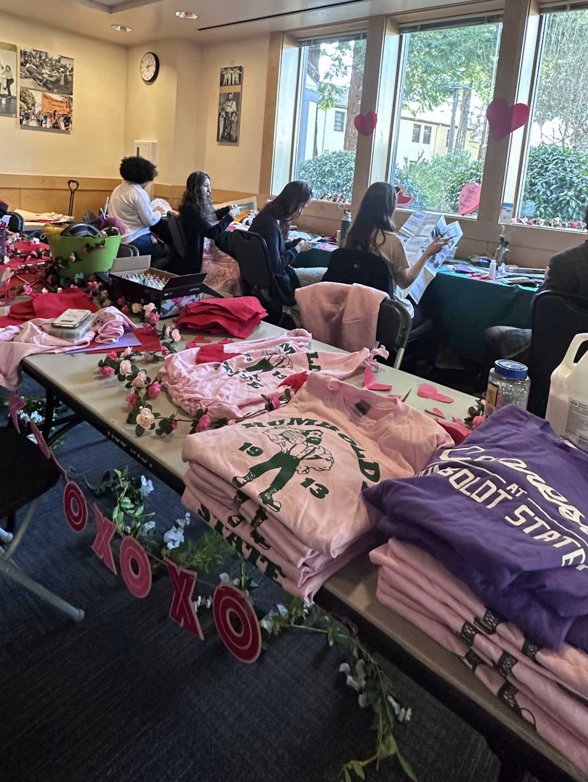 Valentines tea party event image of one of the crafting tables with pink and purple donated vintage Humboldt state t-shirts free to decorate and upcycle , students in the background decorating valentines gifts