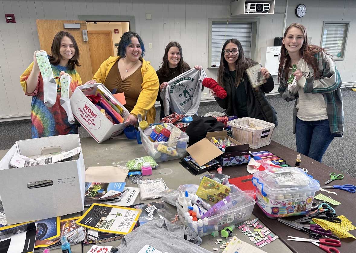 5 EcoReps smiling infront of the crafting table at the DIY day event. Crafting supplies donated and sourced from campus are on the table infront of the cohort members. Some members are holding up socks and HSU t-shirts they upcycled