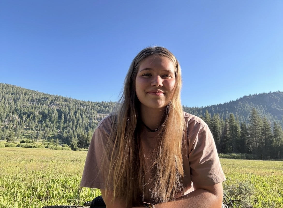 Allison sitting in a sunny, green meadow with a forested, low mountain ridge in the background. The sky is blue and cloudless.