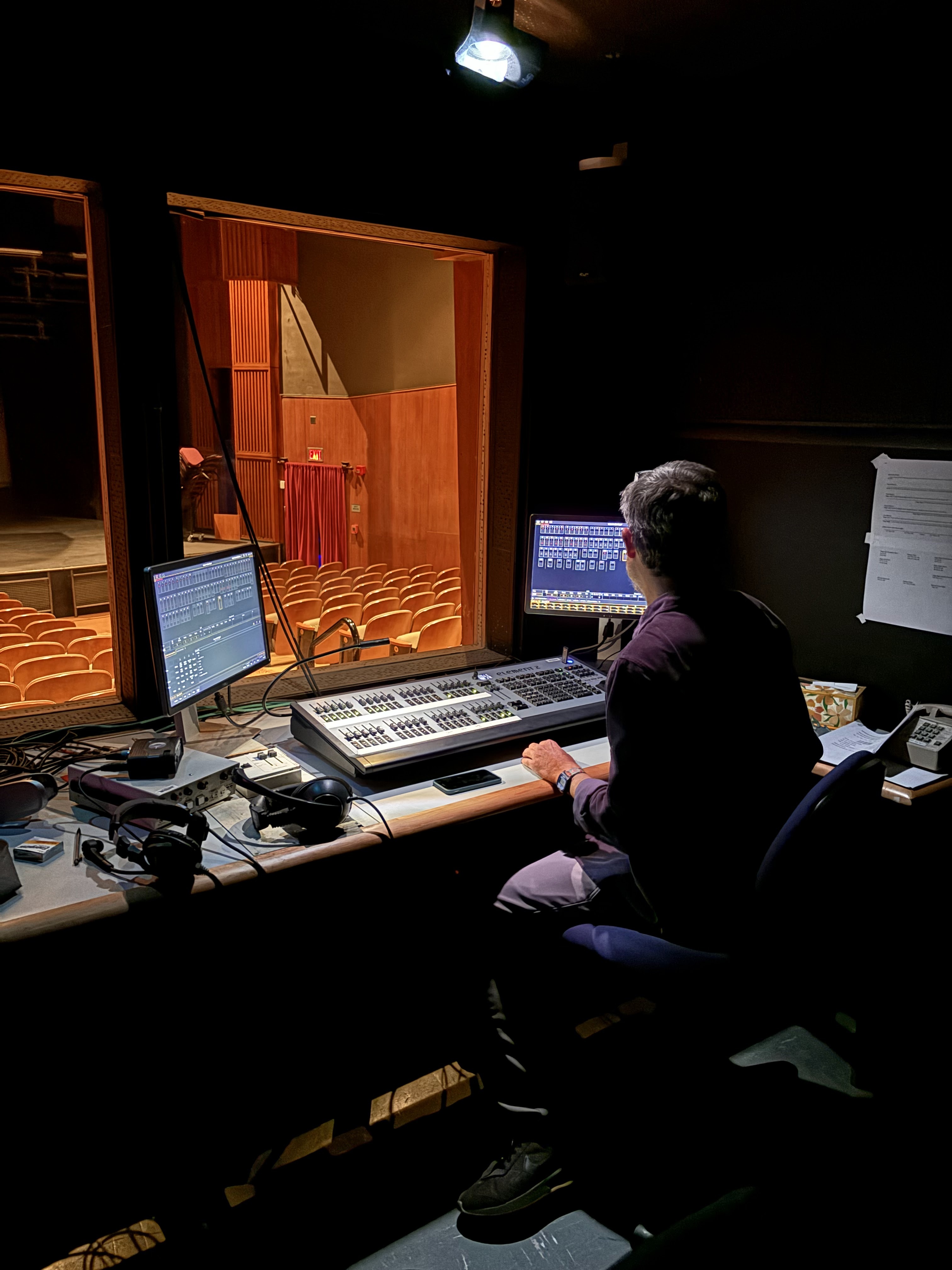 Man in theatre lighting studio with lighting control board in front of him, below two computer screens. 