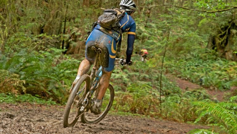 A person on a mountain bike covered in mud riding on a single track trail.