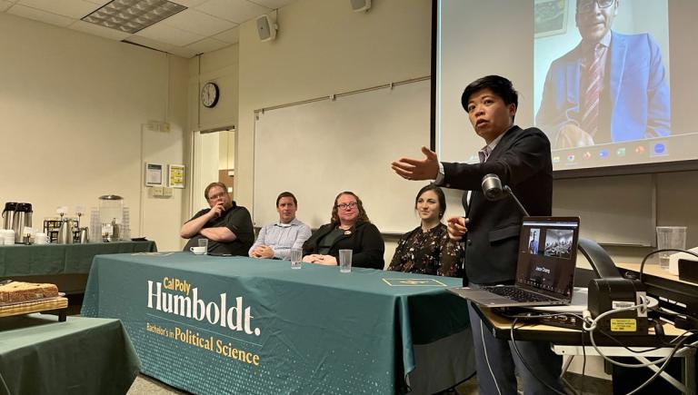 Professor speaking to the room and a table of people behind them