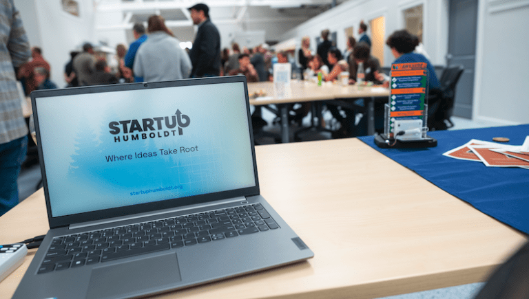 A laptop on a table with StartUp Humboldt logo and people mingling in the background.