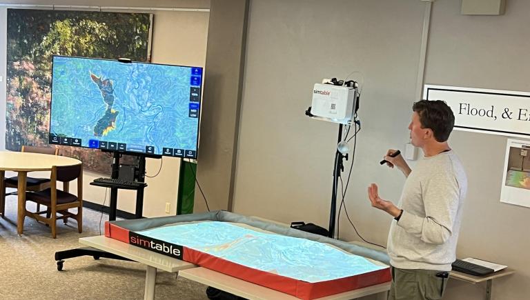 Someone using a laser pointer to affect the Fire SimTable projection in the sand-table and a monitor displaying a map of a fire.