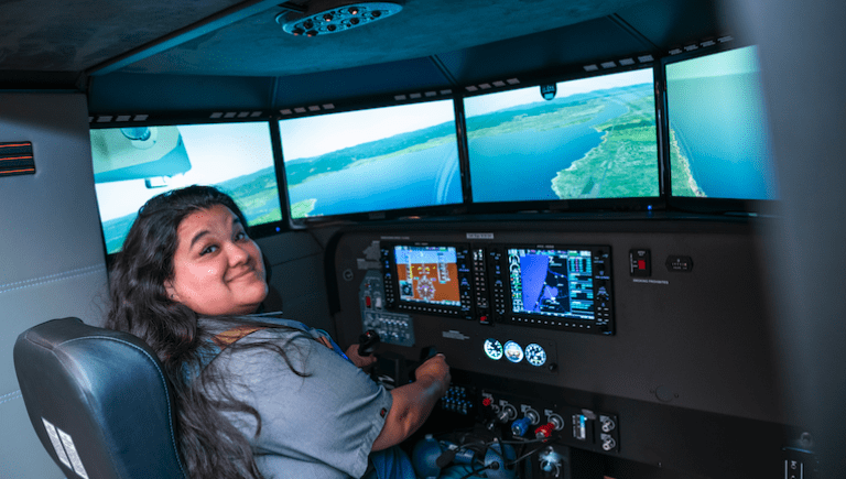A student looking back at the camera in front of a flight simulator.