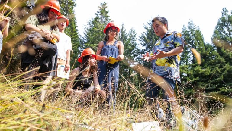 Four students standing in a field wearing orange hard hats listening attentively to a lecture from a faculty member