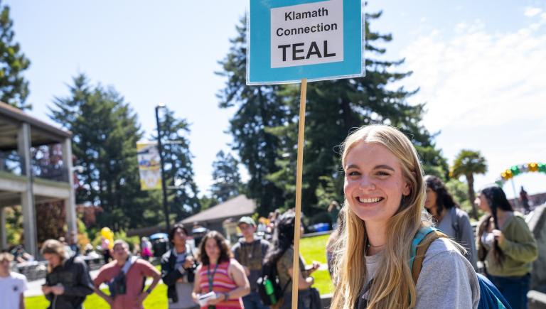 An excited female student holding a blue sign that says "Klamath Connection Teal" in front of a group of students outside on a sunny day