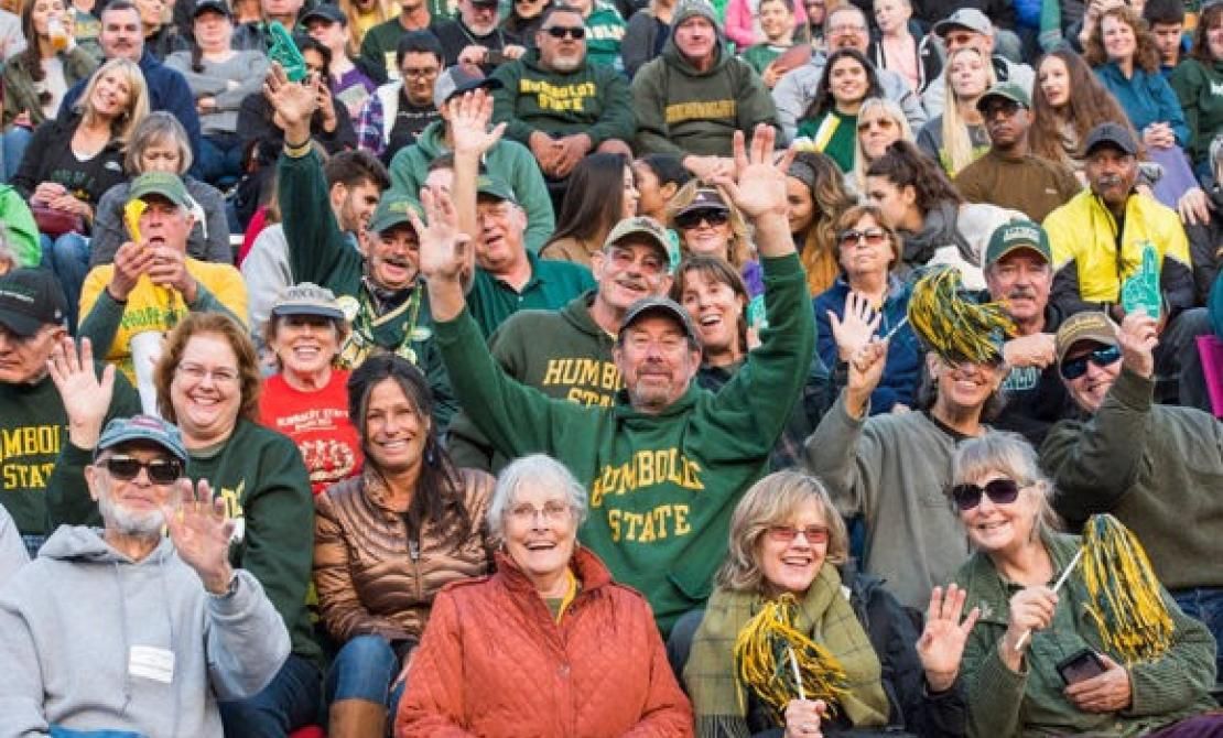 A crowd of enthusiastic Cal Poly Humboldt alumni and community members in the bleachers, many wearing green and gold Humboldt State apparel, smiling and waving at the camera.