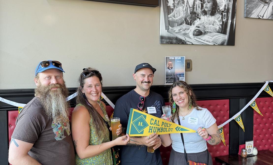 Four Cal Poly Humboldt alumni smile together holding a yellow "Cal Poly Humboldt" pennant at an indoor mixer, with beer glasses in hand and pennant banners decorating the brewery wall behind them.
