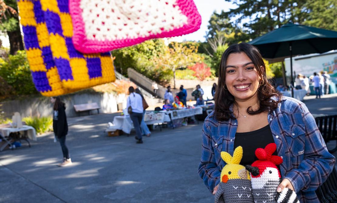 A student with crochet animals at the Makers Market with sales tables in the background.