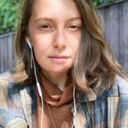 Person wearing a plaid shirt, earbuds, with a fence and greenery in the background