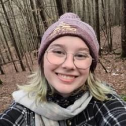 Person wearing a knit cap, plaid shirt, and gray scarf, and glasses. Trees in the background.