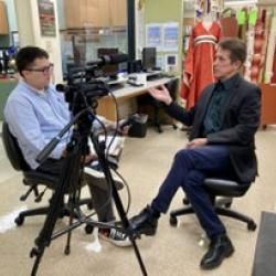 Person interviewing another person. They are sitting in chairs with a camera between them. 