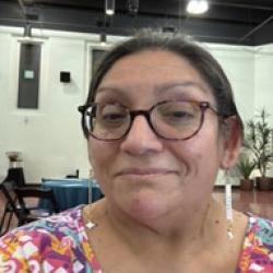 Person wearing a colorful shirt and glasses. An open room with light walls and a dark ceiling in the background.