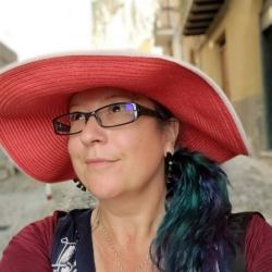 Person wearing a wide-brimmed red hat, glasses, red shirt with black and white vest, outside with a multistory building in the background