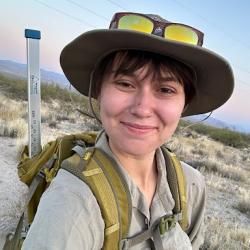 Ayla's portrait, wearing a hat, sunglasses, and a backpack. Out in the wilderness.