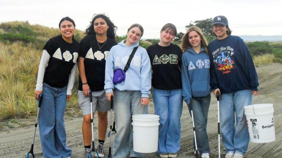Six smiling Delta Phi Epsilon club members with buckets and trash pickers at the beach.