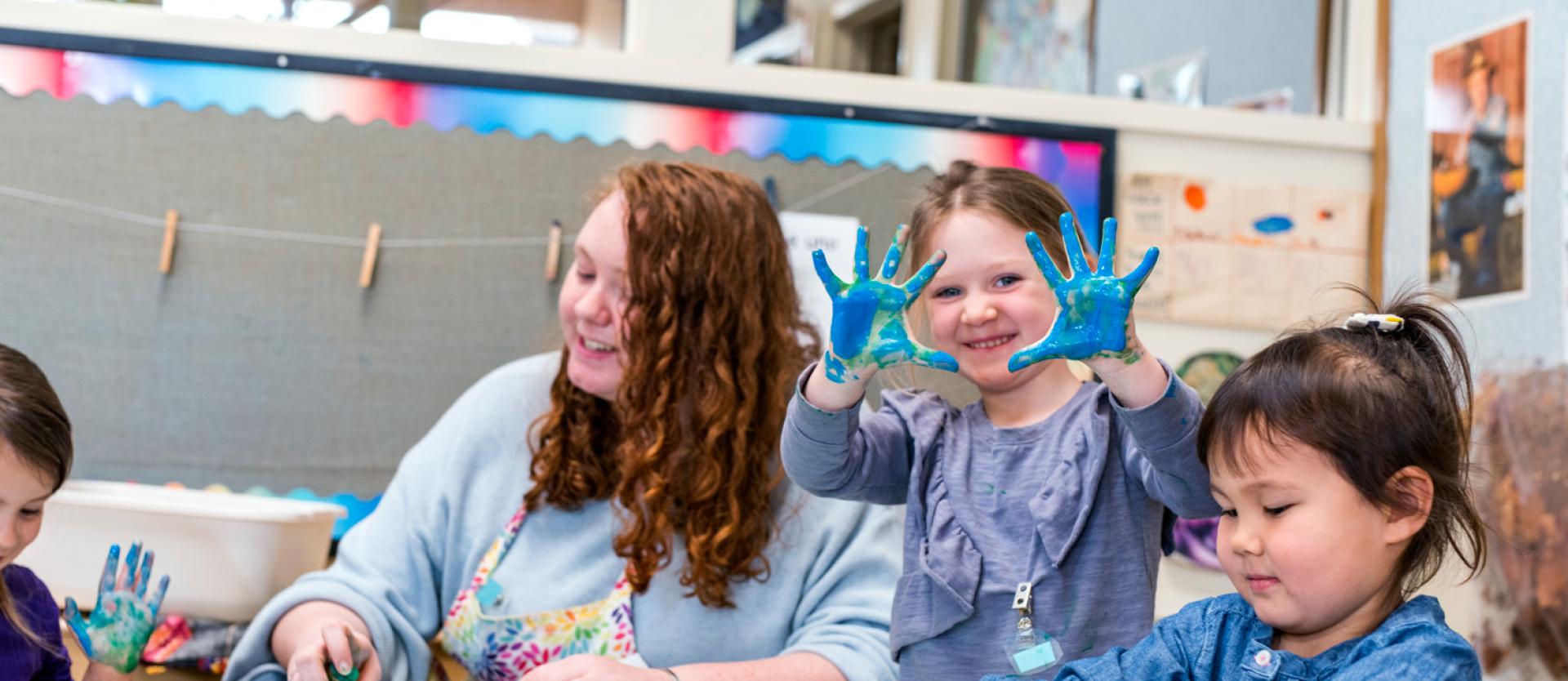 Child holding up her hands with blue paint on them sitting next to a teacher and another child
