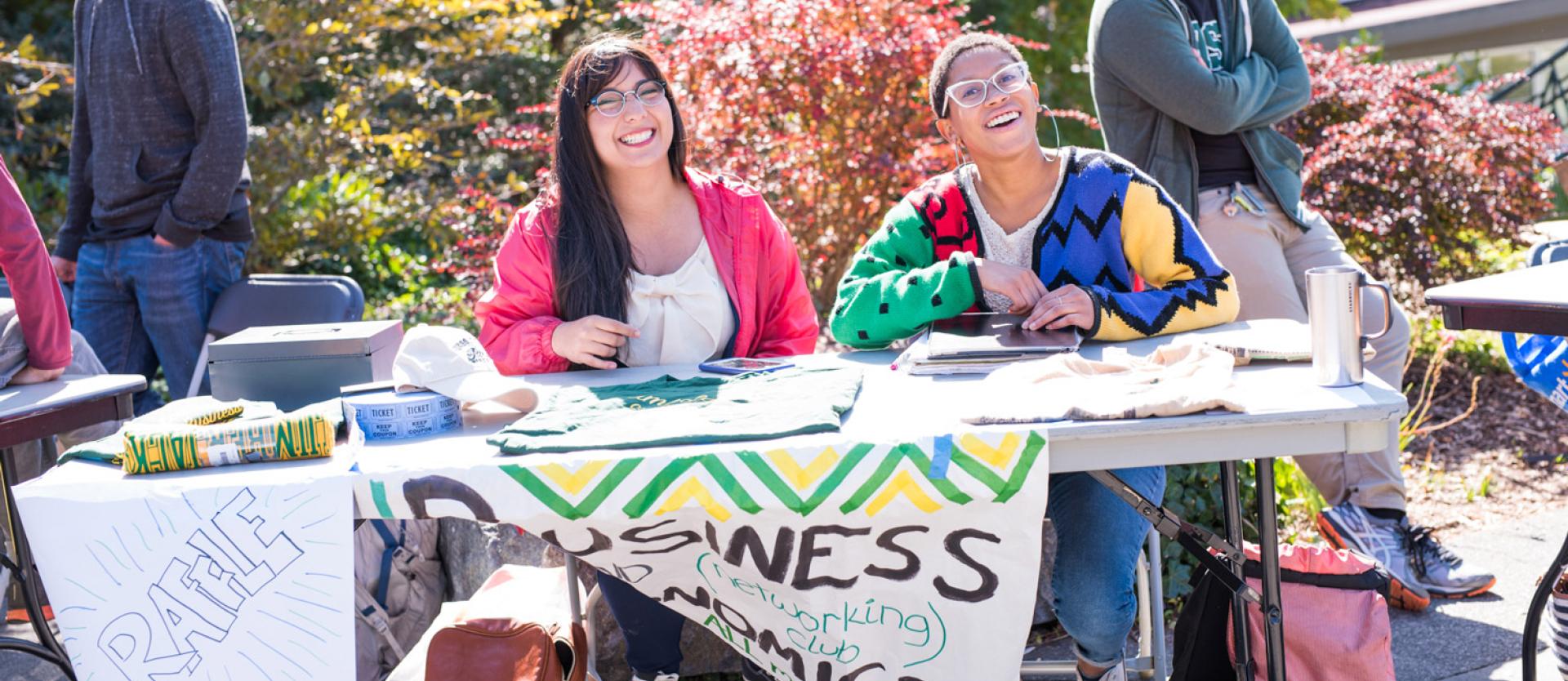 Two students sitting in front of a table outside smiling at the cameral