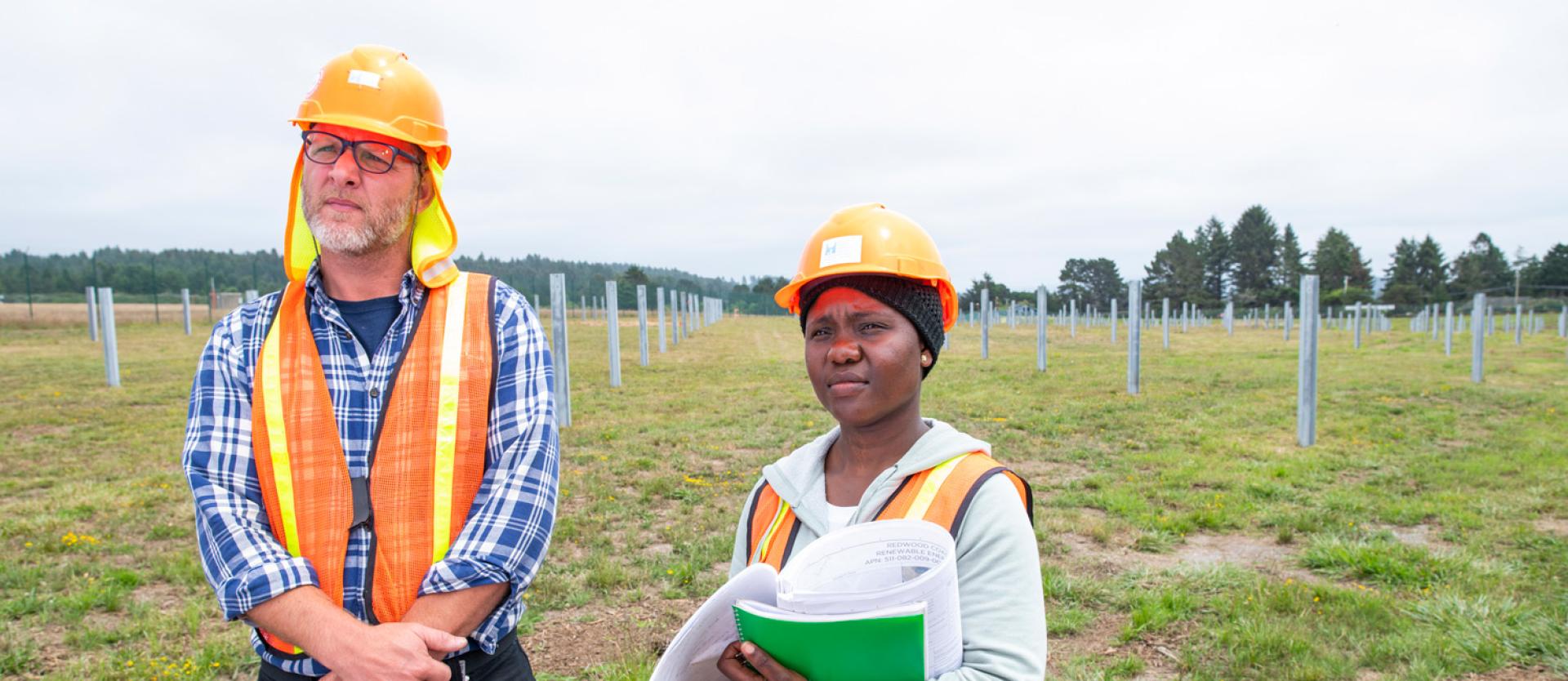 two people in the field wearing vests and hard hats carrying a clipboard