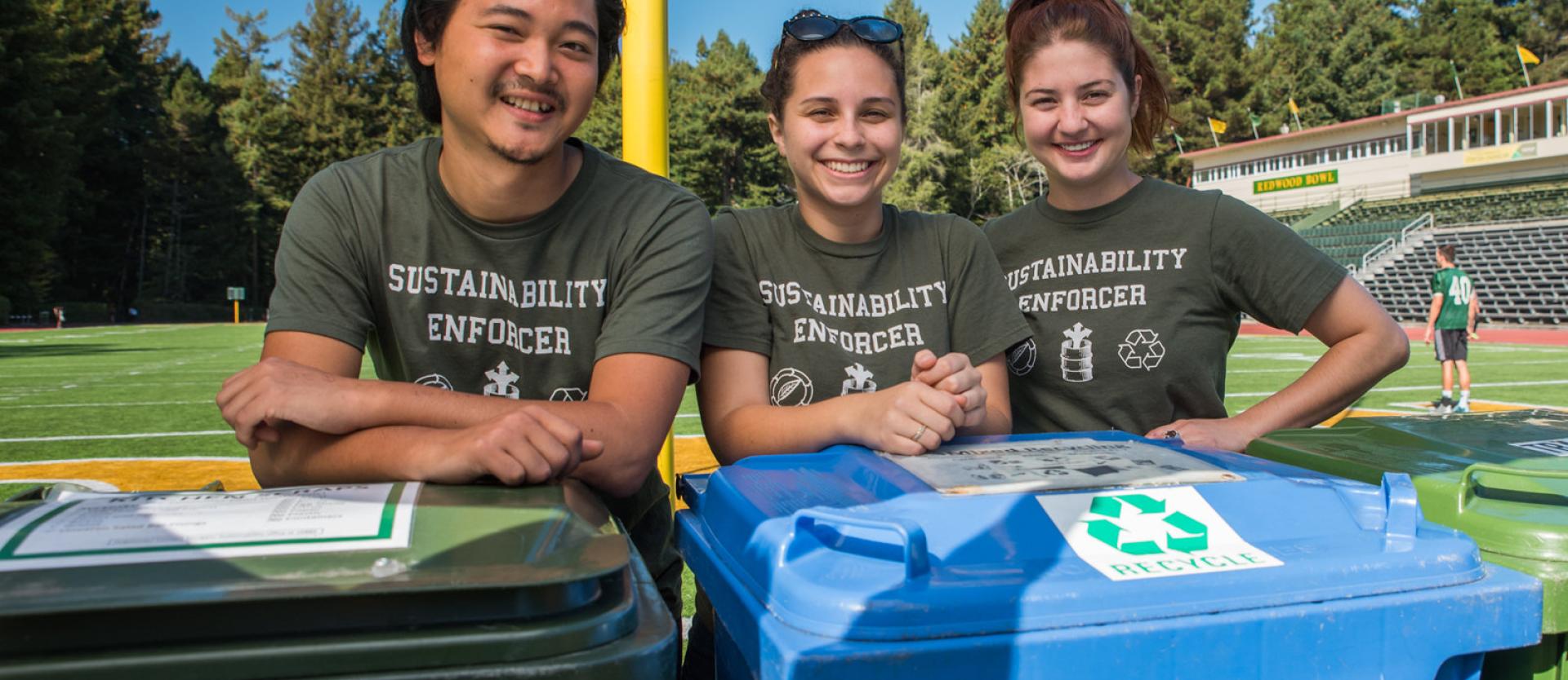Three students wearing shirts that say Sustainability Enforcer standing in front of two recycling bins