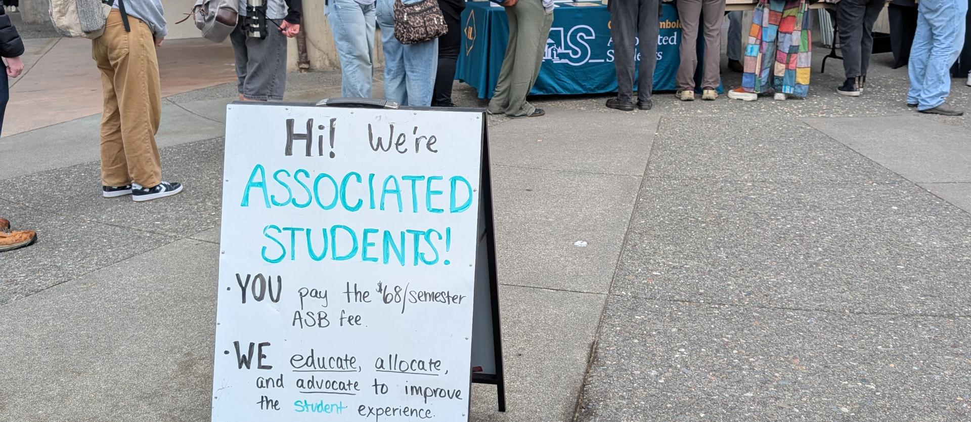 Sign says: Hi! We're Associated Students! You: Pay the $68/semester student fee. We: Educate, allocate, and advocate on behalf of the student population. We have donuts!