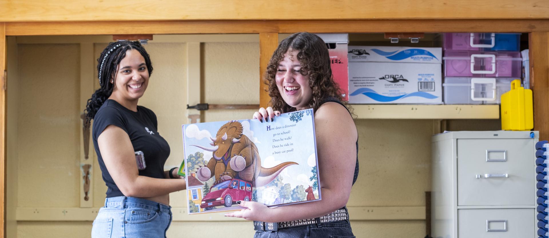 Two college students standing in an elementary school classroom, holding a picture book and smiling.
