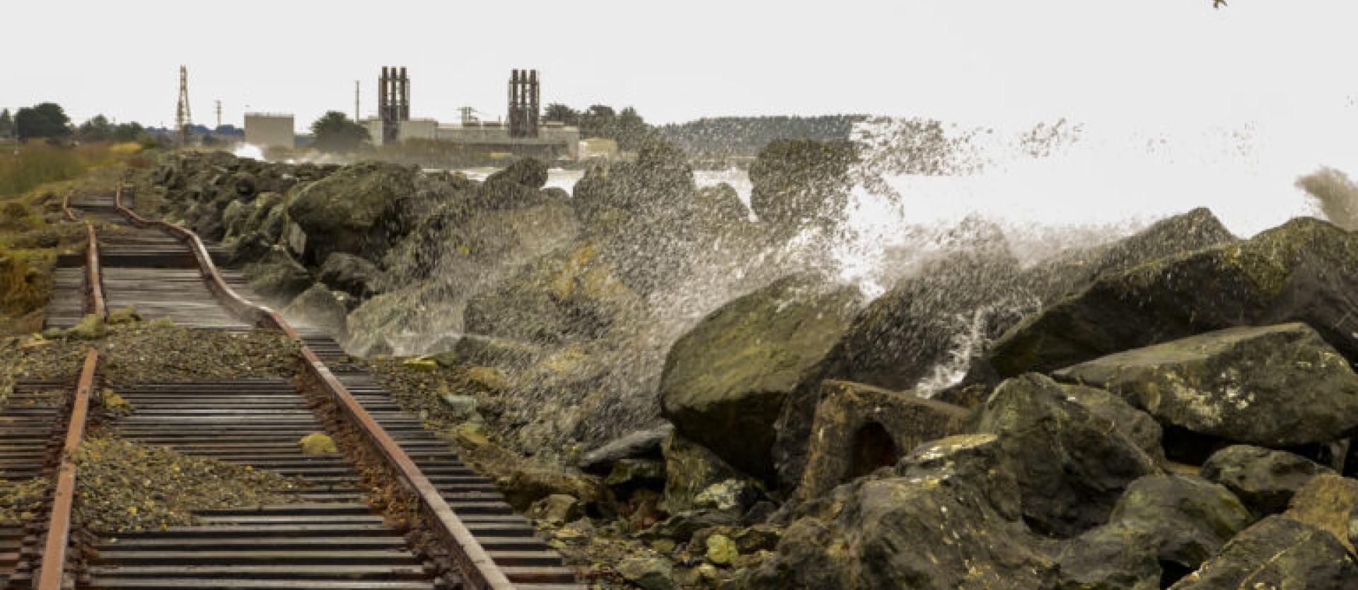 image of waves crashing over a flood wall / rocky rip rap onto a track of rails