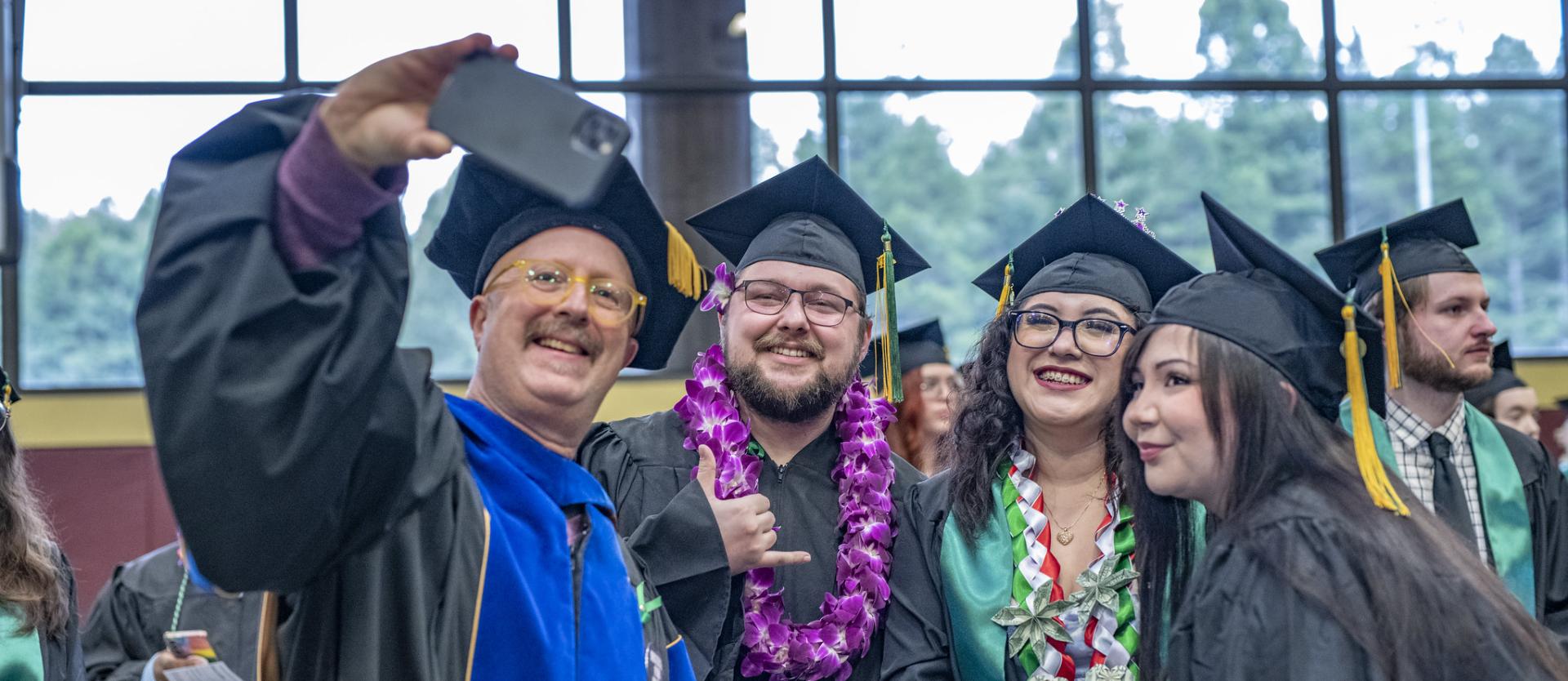 A professor poses for a selfie with three students at graduation ceeremony