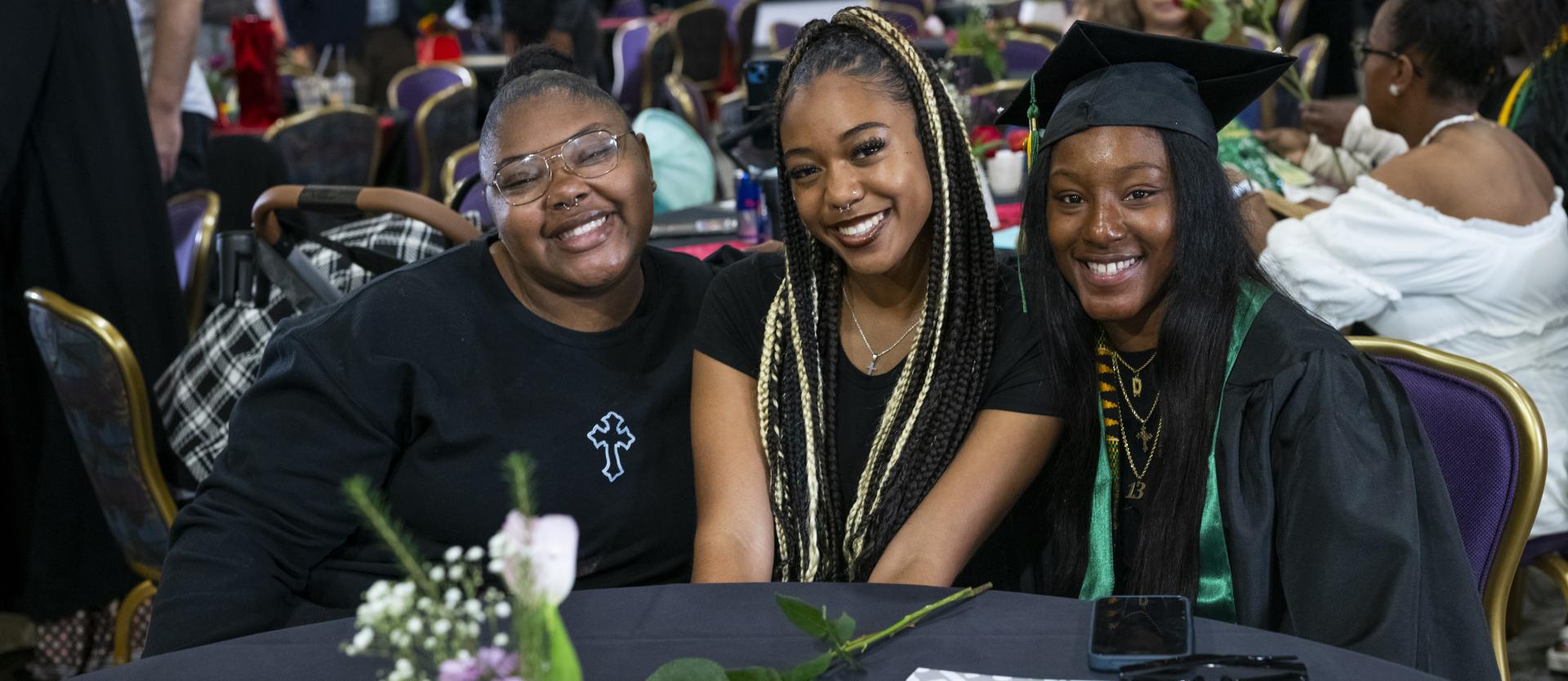 3 students hug and smile together, sitting down at a table during Black Grad 2025