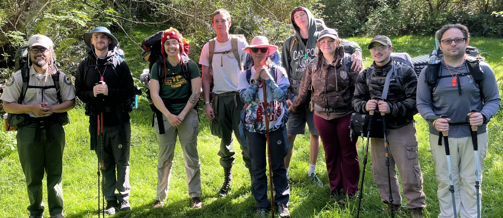 Group photo of backpackers wearing their backpacks in a grassy area surrounded by Trees.
