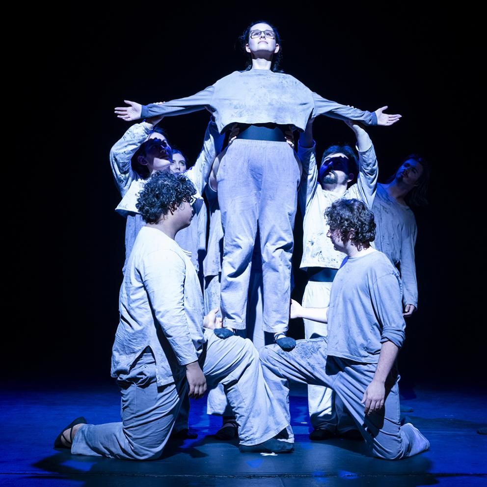 a group of actors dressed in grey holding up another actor
