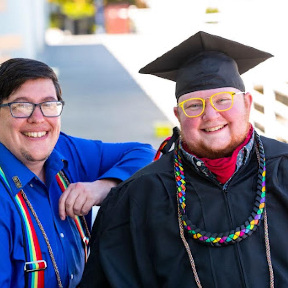 A person in a wheelchair wearing graduation regalia with another person at their side. 