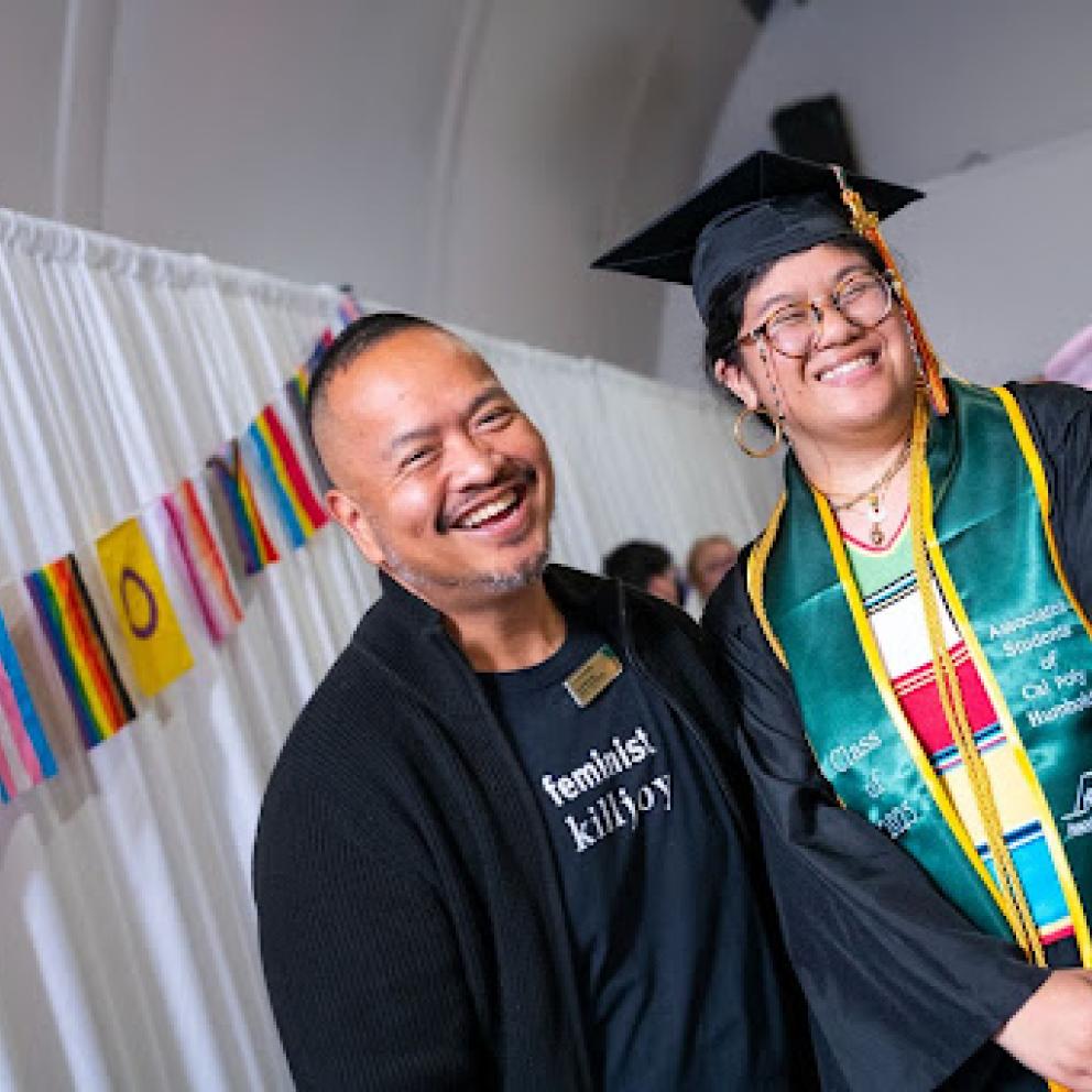 A person with their arm over the shoulder of another person in grad regalia with rainbow flags hanging on the wall.