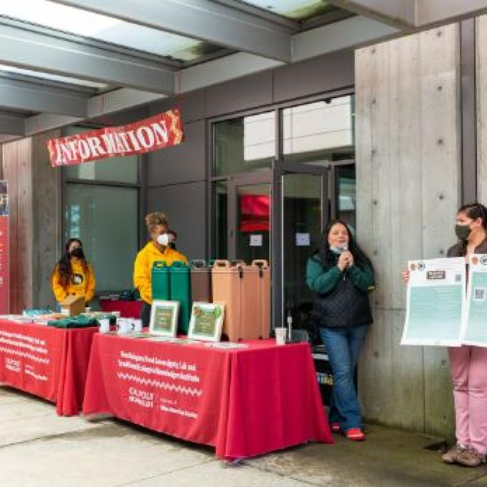 IMage shows members of the Native American Studies Department standing in front of the Rou Dalagurr Food Sovereignty Lab and red table clothed tables with Dr. Cutcha Risling Baldy giving an introduction on the microphone at the first Indigenous Foods Festival in 2023. 