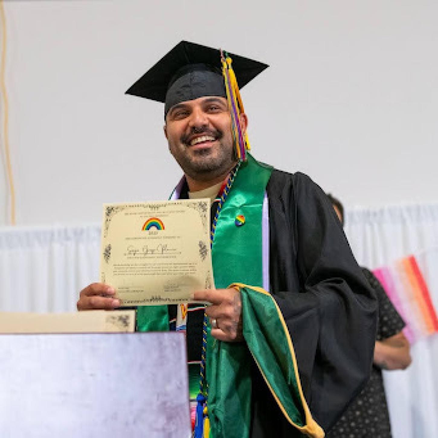 Smiling man in grad regalia holding a rainbow certificate.