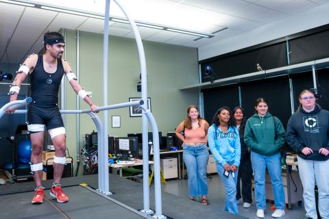 student with electrodes on their body standing on a platform with five other students watching