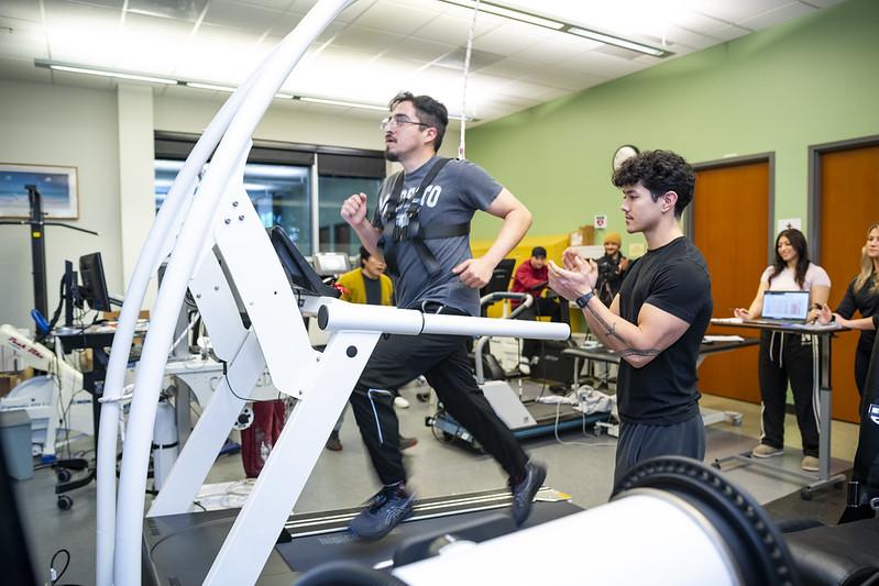 A photo of a student in the Kinesiology Human Performance Lab running on a treadmill wearing a vest that tracks their movement while another student cheers them on. A photo of a student in the Kinesiology Human Performance Lab running on a treadmill wearing a vest that tracks their movement while another student cheers them on.