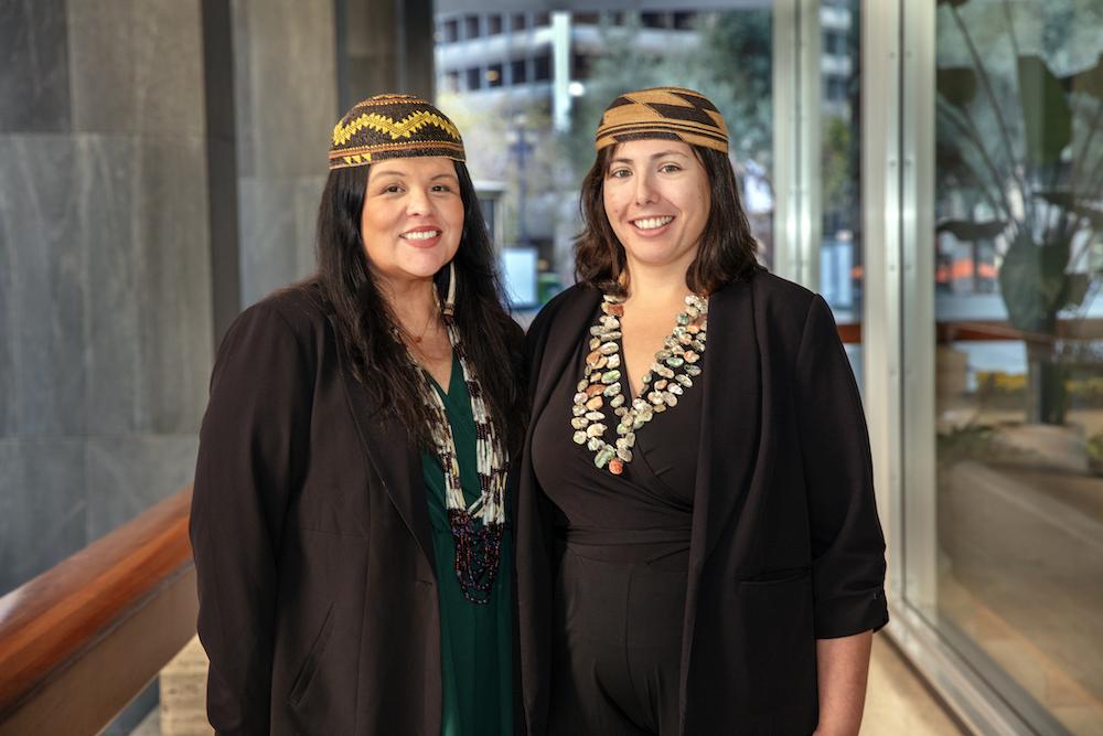 Drs Cutcha Risling Baldy (left) and Kaitlin Reed (right) standing side-by-side smiling in Native dress